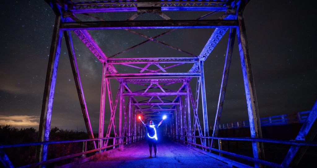 Tukayote Helianthus, standing on the Rio Puerco bridge, wearing lights and illuminating the bridge against the backdrop of the milky way galaxy above.